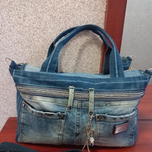 Peut inclure: Sac fourre-tout en jean bleu avec deux poignées et une bandoulière amovible. Le sac est doté d'une fermeture éclair, de poches avant et d'une garniture décorative argentée. Un petit porte-clés est suspendu à la fermeture éclair.