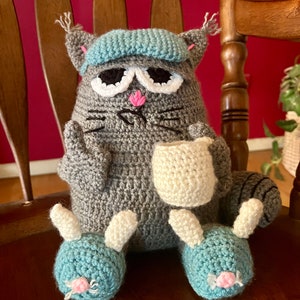 May include: A gray crocheted cat wearing a blue crocheted hat and holding a white crocheted mug. The cat has white eyes and a pink nose. The cat is sitting on a wooden chair with its legs crossed. The cat is wearing blue crocheted slippers with pink noses.