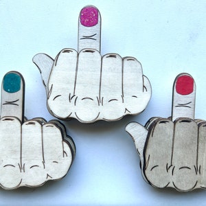 May include: Three wooden hand-shaped ornaments with a middle finger raised. Each ornament has a different colored nail polish on the middle finger: pink, teal, and red.
