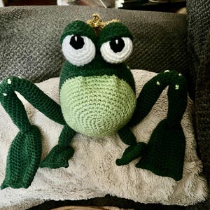 May include: A green crocheted frog wearing a gold crown. The frog has large white eyes with black pupils. The frog is sitting on a light brown fuzzy surface.