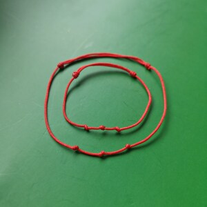 May include: Two red string bracelets with knots, one larger than the other, on a green background.