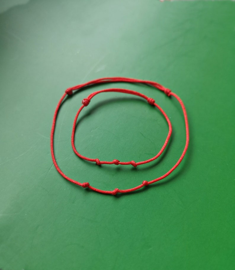 May include: Two red string bracelets with knots, one larger than the other, on a green background.