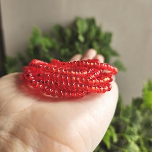 May include: A collection of bright red, translucent beads, likely for jewelry making, are held in a hand. The beads are small and round, strung together to form a flexible strand. The background is blurred with green foliage.