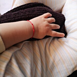May include: A close-up of a person's hand wearing a red string bracelet resting on a brown knitted blanket. The person is wearing a striped shirt.