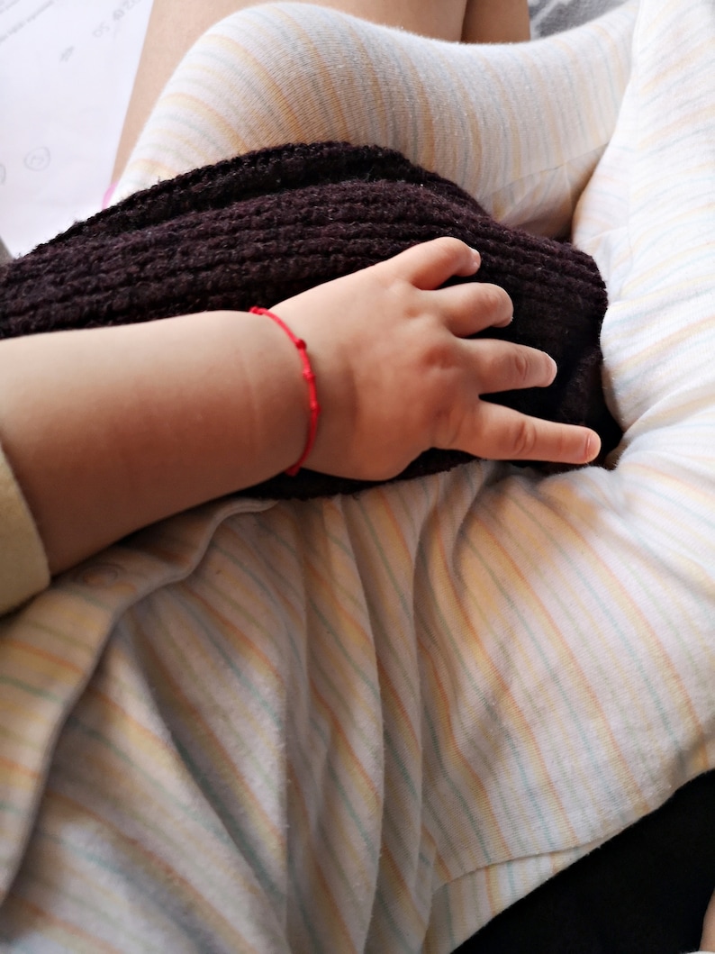 May include: A close-up of a person's hand wearing a red string bracelet resting on a brown knitted blanket. The person is wearing a striped shirt.