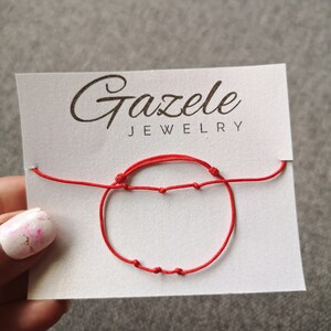 May include: A red string bracelet with small knots and beads. The bracelet is displayed on a white card with the text "Gazelee Jewelry".