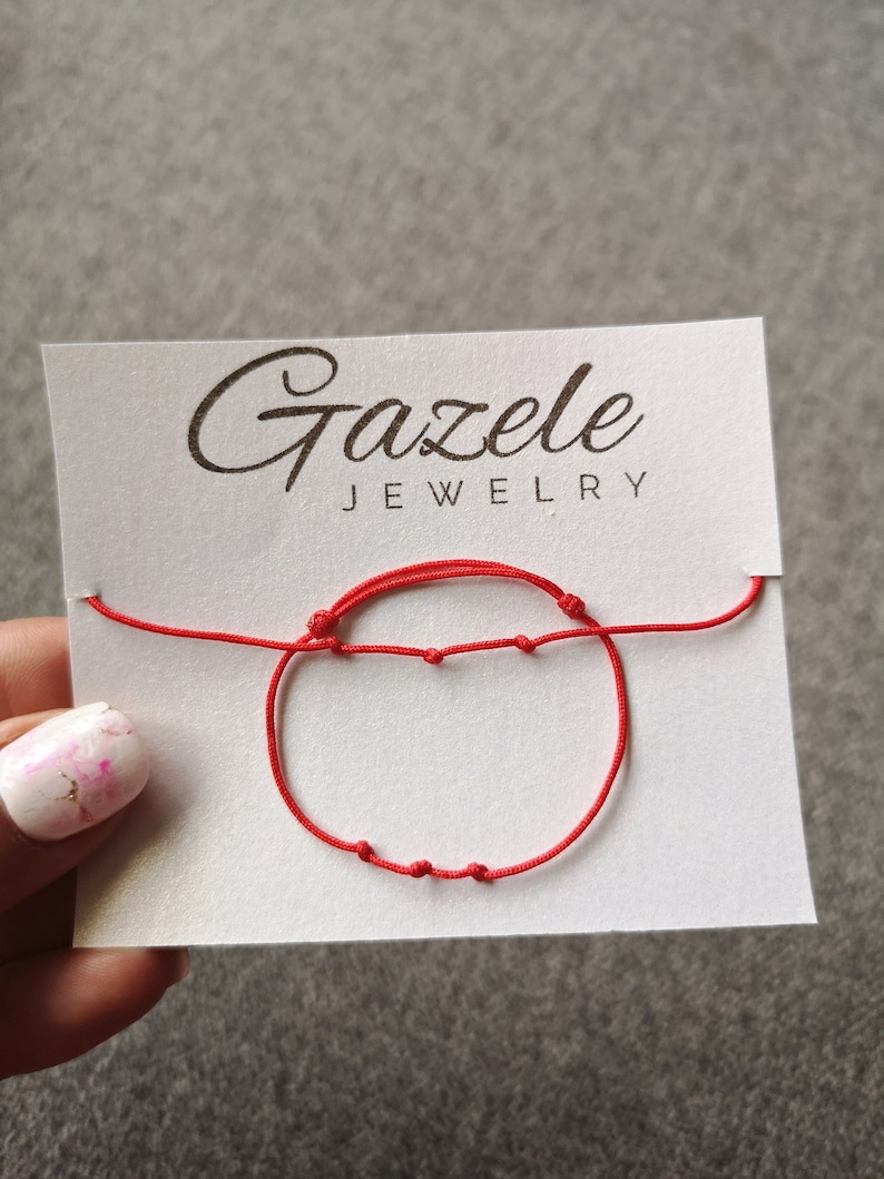 May include: A red string bracelet with small knots and beads. The bracelet is displayed on a white card with the text "Gazelee Jewelry".