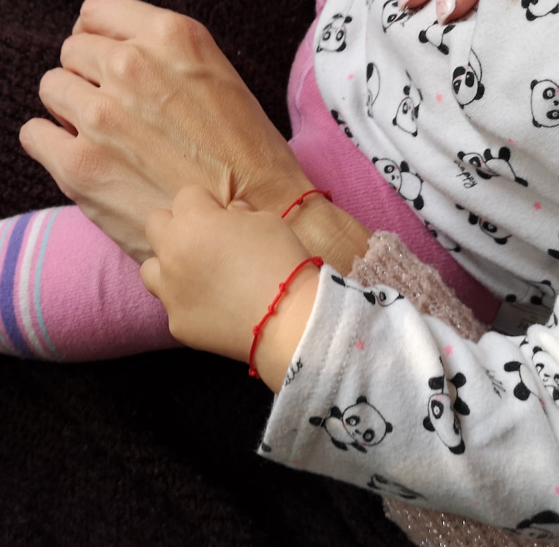May include: Two people wearing red string bracelets on their wrists. The person on the left is wearing a pink and white striped sock. The person on the right is wearing a white shirt with black panda bears printed on it.