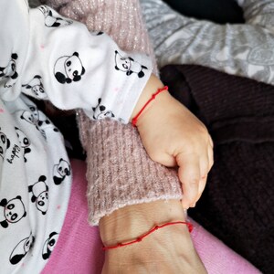 May include: Two people wearing red string bracelets on their wrists. The person on the left is wearing a white shirt with black panda bears printed on it. The person on the right is wearing a pink sweater.