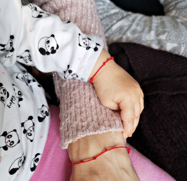 May include: Two people wearing red string bracelets on their wrists. The person on the left is wearing a white shirt with black panda bears printed on it. The person on the right is wearing a pink sweater.