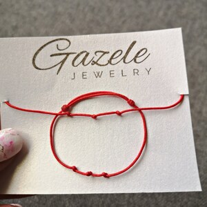 May include: A red string bracelet with knots on a white card with the text "Gazelee Jewelry".