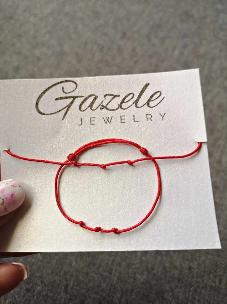 May include: A red string bracelet with knots on a white card with the text "Gazelee Jewelry".
