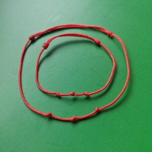 May include: Two red string bracelets with knots, one larger than the other, on a green background.