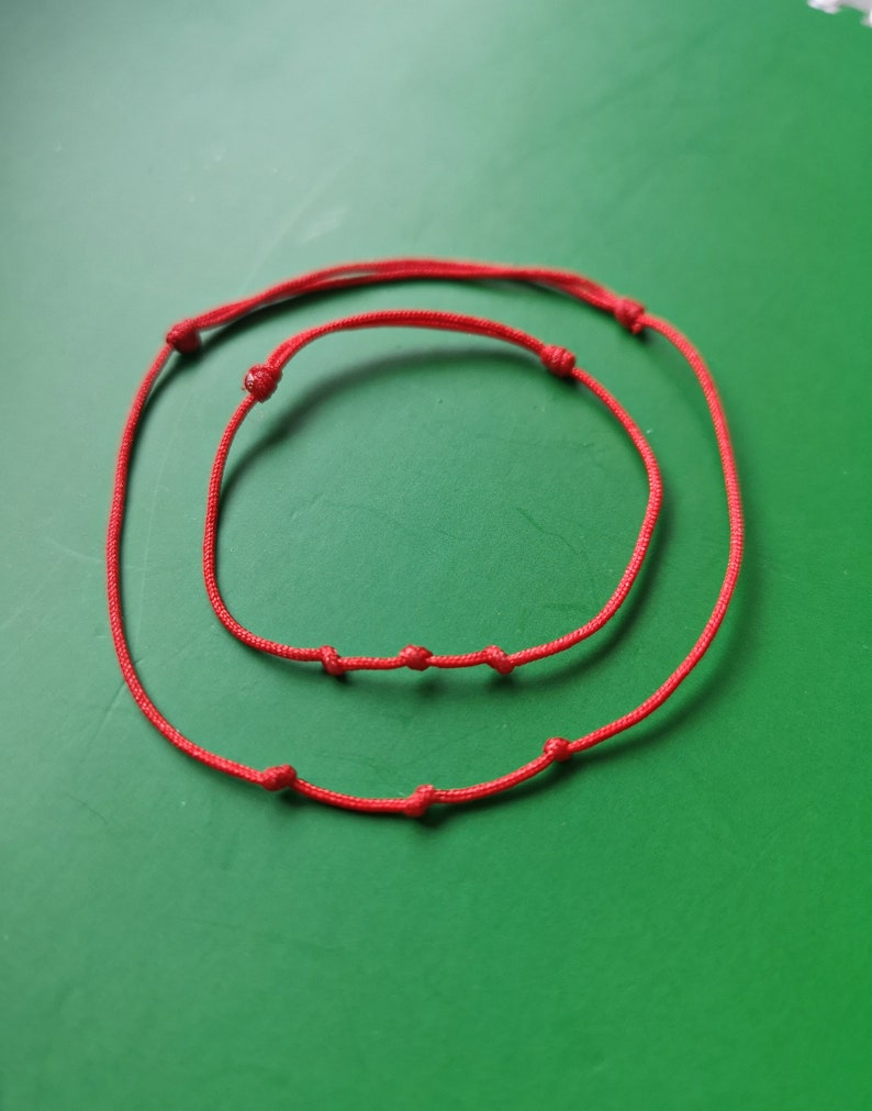 May include: Two red string bracelets with knots, one larger than the other, on a green background.