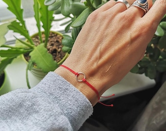May include: A red string bracelet with a small silver circle charm. The bracelet is tied on the wrist. The background includes green plants and a gray long-sleeved shirt.