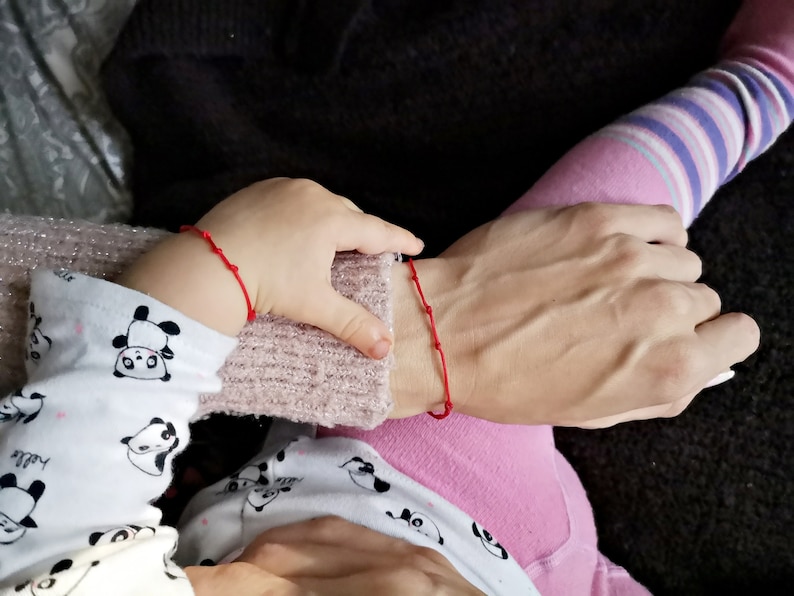 May include: Two people wearing red string bracelets on their wrists. The person on the left is wearing a white shirt with panda bears printed on it. The person on the right is wearing a pink shirt with a striped pattern.