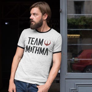 May include: A light gray ringer t-shirt with black trim and the text "TEAM MOTHMA" in black, with a red graphic. The person is wearing blue jeans. The shirt is made of a soft, comfortable material.