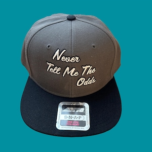 May include: Gray and black baseball cap with the text "Never Tell Me The Odds" embroidered in white on the front. The cap has a black snap closure and a silver label with the text "S-N-A-P" and "The Premium Snapback".