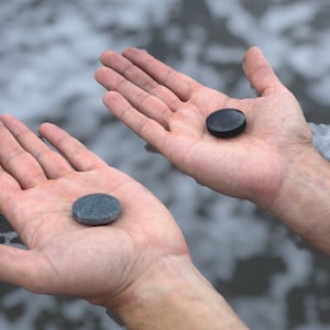 Peut inclure: Deux pierres lisses et rondes, l'une grise et l'autre noire, sont tenues dans des mains ouvertes sur un fond flou d'eau. Les pierres semblent naturelles, peut-être ramassées sur une plage ou au bord d'une rivière. L'image se concentre sur les pierres et la texture des mains.