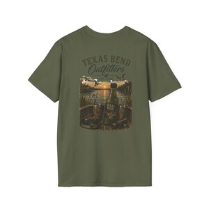 May include: An olive green t-shirt featuring a graphic print of a sunset over a lake with a bottle labeled Texas Bend, two metal cups, and flying ducks. The text reads Texas Bend Outfitters Est 2024, with Texas Bend on the bottle label.