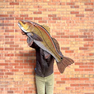May include: A large, detailed fish sculpture with a red, yellow, and brown color scheme. The artwork features intricate scale and fin details, and is held up against a brick wall. The fish is a decorative item.