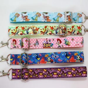 Könnte beinhalten: Fünf farbenfrohe Armbandschlaufen mit silberfarbenen Verschlüssen. Die Schlaufen zeigen Zeichentrickfiguren auf hellblauem, grünem, rosa, blauem und lila Hintergrund. Auf dem rosa Band steht das Wort "TOY".