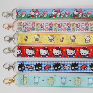 May include: Six fabric wristlet keychains featuring Sanrio character prints. The top strap is light blue with various characters, followed by a pink strap with multiple icons, a yellow Hello Kitty strap, a red Hello Kitty strap, a blue gingham strap with Badtz-Maru and sushi, and a light teal strap with Pompompurin.