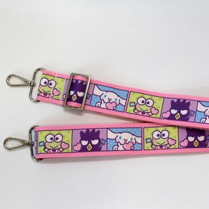 May include: A pink and blue patterned strap with cartoon character illustrations. The strap has silver metal clasps and a buckle. The design features repeating squares with characters on a pink background.