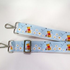 May include: Light blue strap with a repeating pattern of a yellow bear, white daisies, and bees. The strap has silver metal clips and a bronze-colored metal buckle. The strap is designed for a bag or purse.