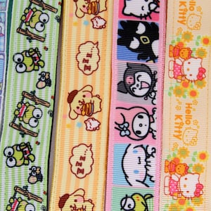 Peut inclure: Quatre bandes de ruban gros-grain avec des personnages Sanrio. La première est vert clair avec Keroppi sur des balançoires. La deuxième est jaune avec Pompompurin et des bulles de texte ZZZ. La troisième a des carrés colorés avec Kuromi, My Melody et Cinnamoroll. La quatrième est pêche avec Hello Kitty et des tournesols.