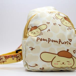 May include: A cream-colored sling bag featuring a repeating pattern of light brown shapes and cartoon character illustrations. The bag has a yellow zipper and strap, with the words "Pompompurin" printed on the front.