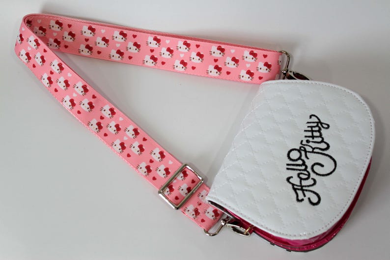 Puede incluir: Un bolso bandolera rosa con una correa a juego con un patr&oacute;n repetido de caras y corazones de Hello Kitty. El bolso tiene una parte delantera acolchada blanca con "Hello Kitty" en escritura negra.