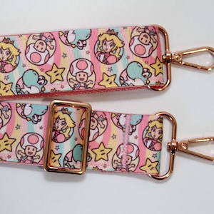 May include: A pink and pastel-colored bag strap featuring a repeating pattern of Princess Peach, Toad, Yoshi, and stars. The strap has gold-colored metal hardware, including a buckle and two swivel clasps.