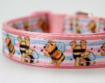 May include: A pink dog collar with a repeating pattern of cartoon bees and Winnie the Pooh characters. The collar has a light pink trim and a metal clasp. The background of the pattern has pink, blue, and white stripes.