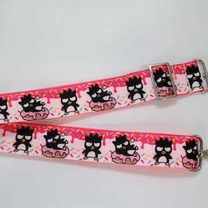 May include: A pink and white patterned strap featuring cartoon penguins on donuts. The strap has silver metal hardware and is designed for a bag or purse. The strap is approximately 1 inch wide.