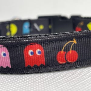 May include: Black dog collar with a Pac-Man themed design. The collar features pink and red ghosts, cherries, and a yellow Pac-Man.
