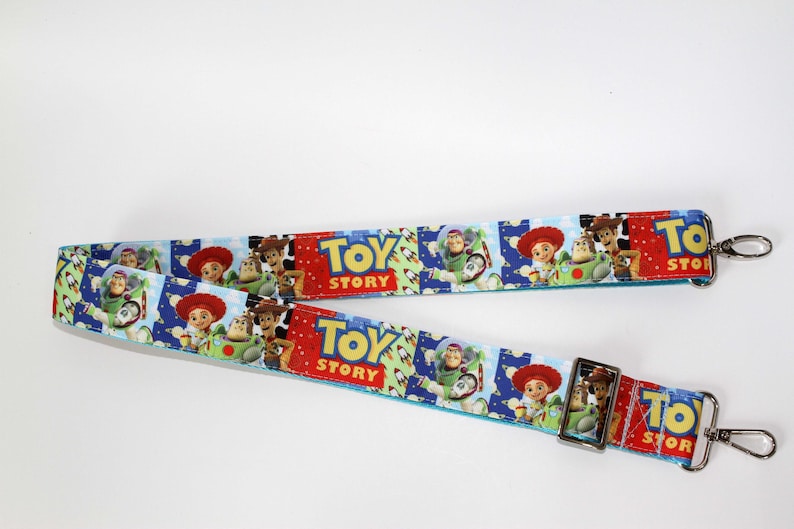 Puede incluir: Una correa azul y blanca con tem&aacute;tica de Toy Story con im&aacute;genes de Woody, Buzz Lightyear y Jessie. La correa tiene cierres met&aacute;licos plateados y las palabras "Toy Story" en rojo y amarillo.