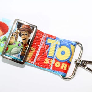 Puede incluir: Un cord&oacute;n con personajes de la pel&iacute;cula de animaci&oacute;n "Toy Story". El cord&oacute;n tiene un fondo azul con nubes blancas y elementos espaciales. Incluye im&aacute;genes de Jessie, Woody y Buzz Lightyear, con las palabras "Toy Story" en amarillo.