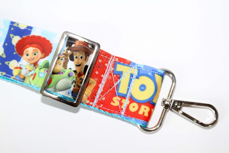 Puede incluir: Un cord&oacute;n con personajes de la pel&iacute;cula de animaci&oacute;n "Toy Story". El cord&oacute;n tiene un fondo azul con nubes blancas y elementos espaciales. Incluye im&aacute;genes de Jessie, Woody y Buzz Lightyear, con las palabras "Toy Story" en amarillo.