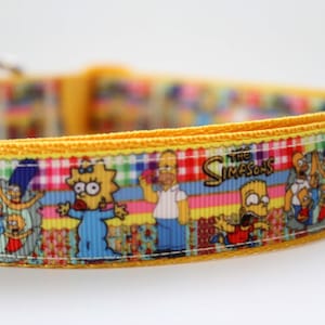 Op de afbeelding: Een gele halsband voor huisdieren met een herhalend patroon van The Simpsons-personages, waaronder Homer, Marge, Bart, Lisa en Maggie. Het ontwerp bevat het logo van de serie en een kleurrijke ruitjesachtergrond met een gele kunststof gesp.