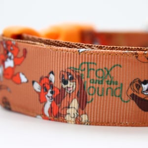 May include: Brown dog collar featuring illustrations of Tod the fox and Copper the hound, with the text "Fox and the Hound" in green. The collar has an orange buckle and is made of a durable, ribbed material.