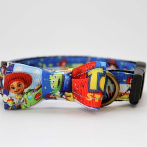 May include: A blue dog collar with a black buckle and a bow tie featuring characters from the animated film Toy Story. The bow tie has a red, yellow, and blue design.