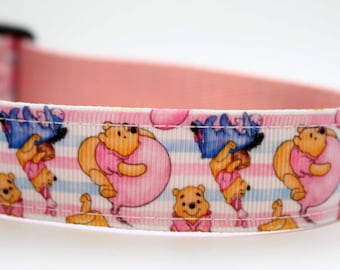 May include: A dog collar with a repeating pattern of Winnie the Pooh characters. The design features Pooh Bear, Eeyore, and other characters on a striped background. The collar has a pink interior and is primarily white and pink.