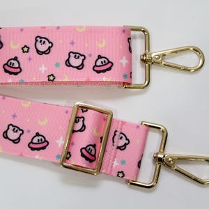 Puede incluir: Una correa de bolso ajustable de color rosa con un patrón repetitivo de personajes de dibujos animados, pequeños platillos voladores, lunas crecientes amarillas y estrellas multicolores. Incluye herrajes de metal dorado y dos mosquetones.