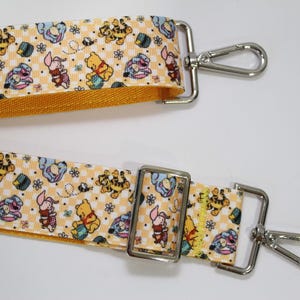 Puede incluir: Una correa estampada con personajes de dibujos animados, con Winnie the Pooh, Piglet, Tigger y Eeyore. La correa tiene un fondo a cuadros amarillo y blanco y herrajes de metal plateado. Probablemente para un bolso o cartera.