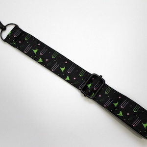 May include: Black adjustable strap with a repeating pattern of green witch hats, pink stars, and pink potion bottles. The strap has a black metal swivel clasp and a black metal slider buckle. The strap is likely for a bag or purse.