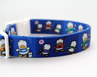 Pode incluir: Uma coleira azul para animais de estimação com um padrão repetitivo de patos de desenho animado vestindo várias camisas coloridas. A coleira inclui uma fivela de plástico branco, um ajustador de plástico branco e um anel em D de metal prateado.