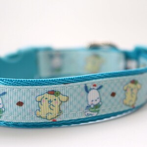 May include: A light blue pet collar featuring a repeating pattern of cartoon characters, including yellow and white figures with small red accents. The collar is constructed from woven fabric with a matching blue plastic buckle and hardware.