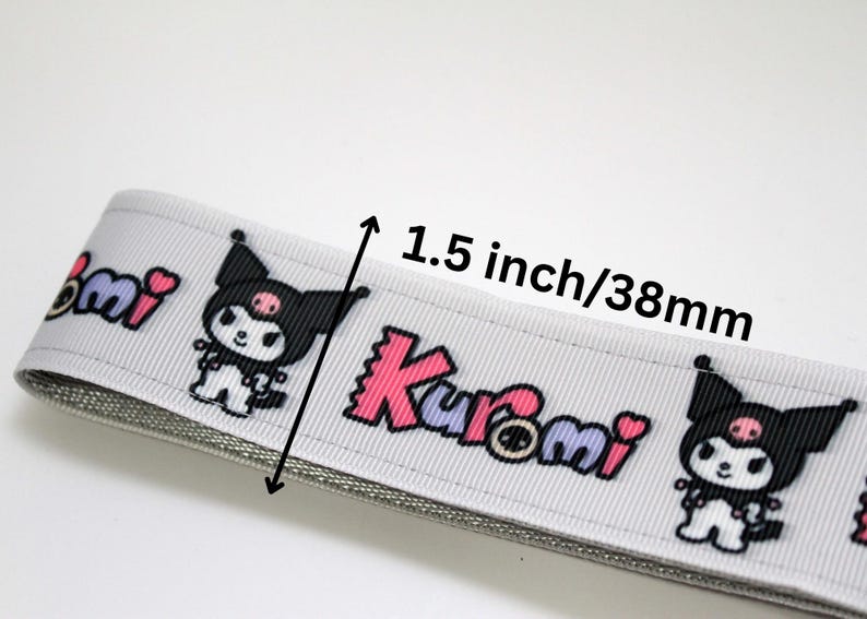 Puede incluir: Cinta gris claro con un patr&oacute;n repetido de un personaje de dibujos animados y la palabra "Kuromi" en rosa y morado. La cinta mide 3,8 cm de ancho.