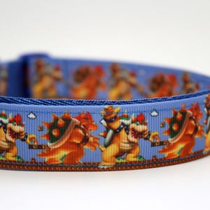 May include: A blue dog collar featuring a repeating pattern of the Bowser character from the Super Mario video game series. The collar has a blue background with Bowser in orange, yellow, and green. The collar is made of a durable material.